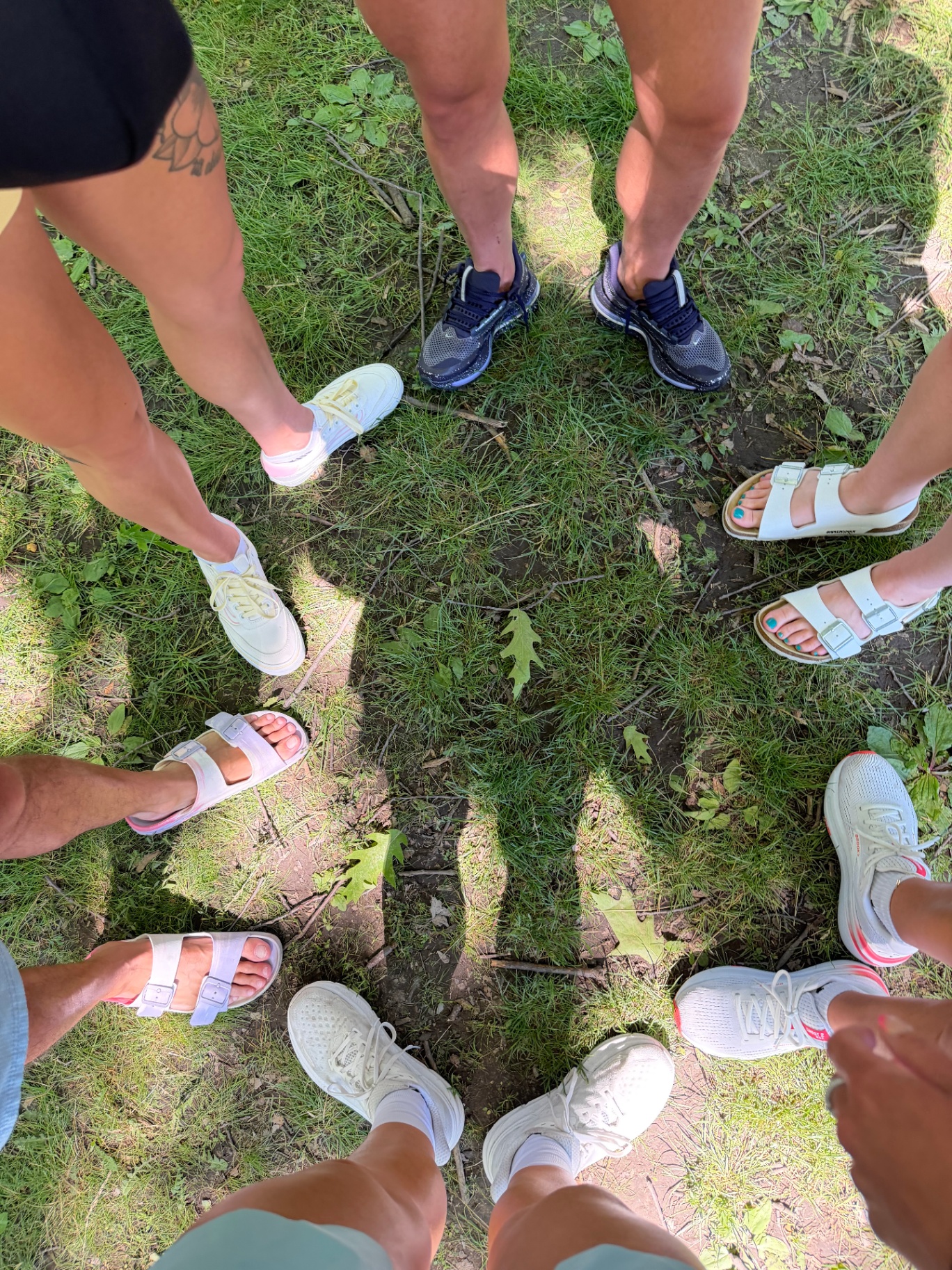 A circle of friends standing together on grass, seen from above — feet and shoes in a ring of community and connection